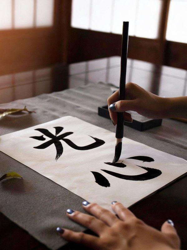 Photo showing a hand holding a calligraphy brush drawing the Japanese kanji for Ko Shin - the path of light - on a piece of natural paper.