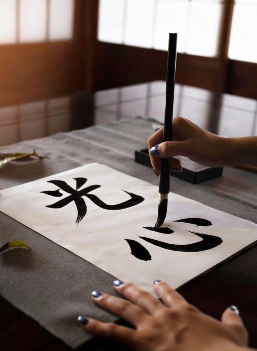 Photo showing a hand holding a calligraphy brush drawing the Japanese kanji for Ko Shin - the path of light - on a piece of natural paper.